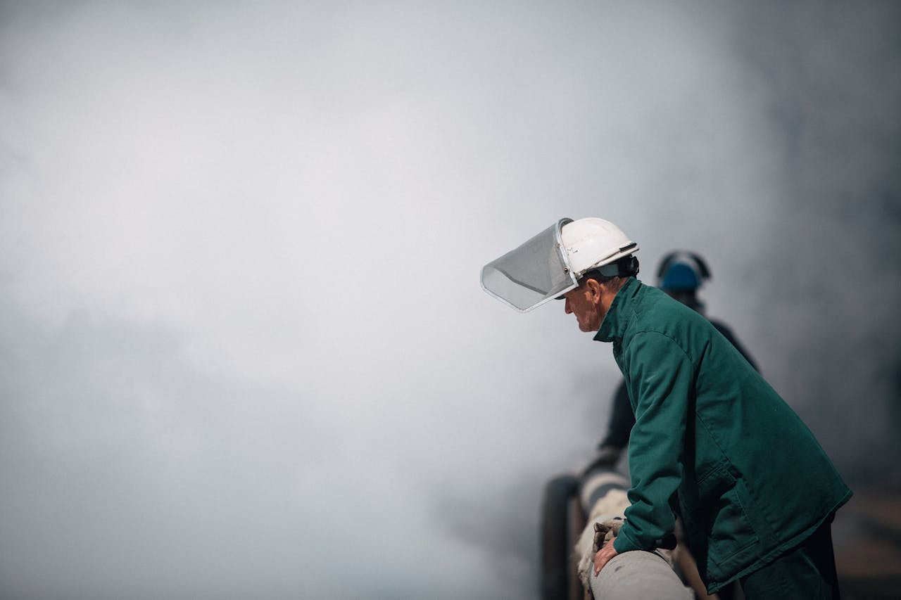 Industrial worker in protective gear inspecting a large pipe amidst dense smoke in an outdoor setting.