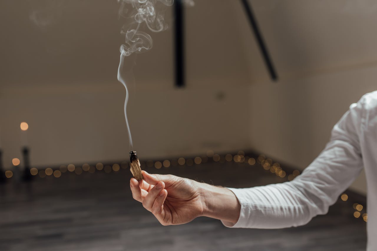 Close-up of a hand holding a smoldering Palo Santo stick, smoke curling in a calm indoor setting.